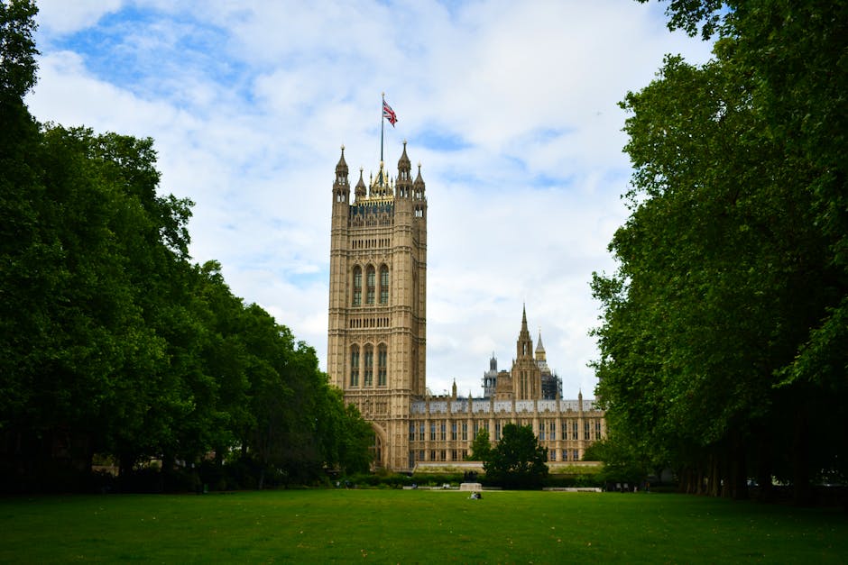 A historic Gothic-style tower with ornate spires and detailed stonework, prominently featuring the Union Jack flag flying from its highest point, set against a partly cloudy sky. The tower is surround